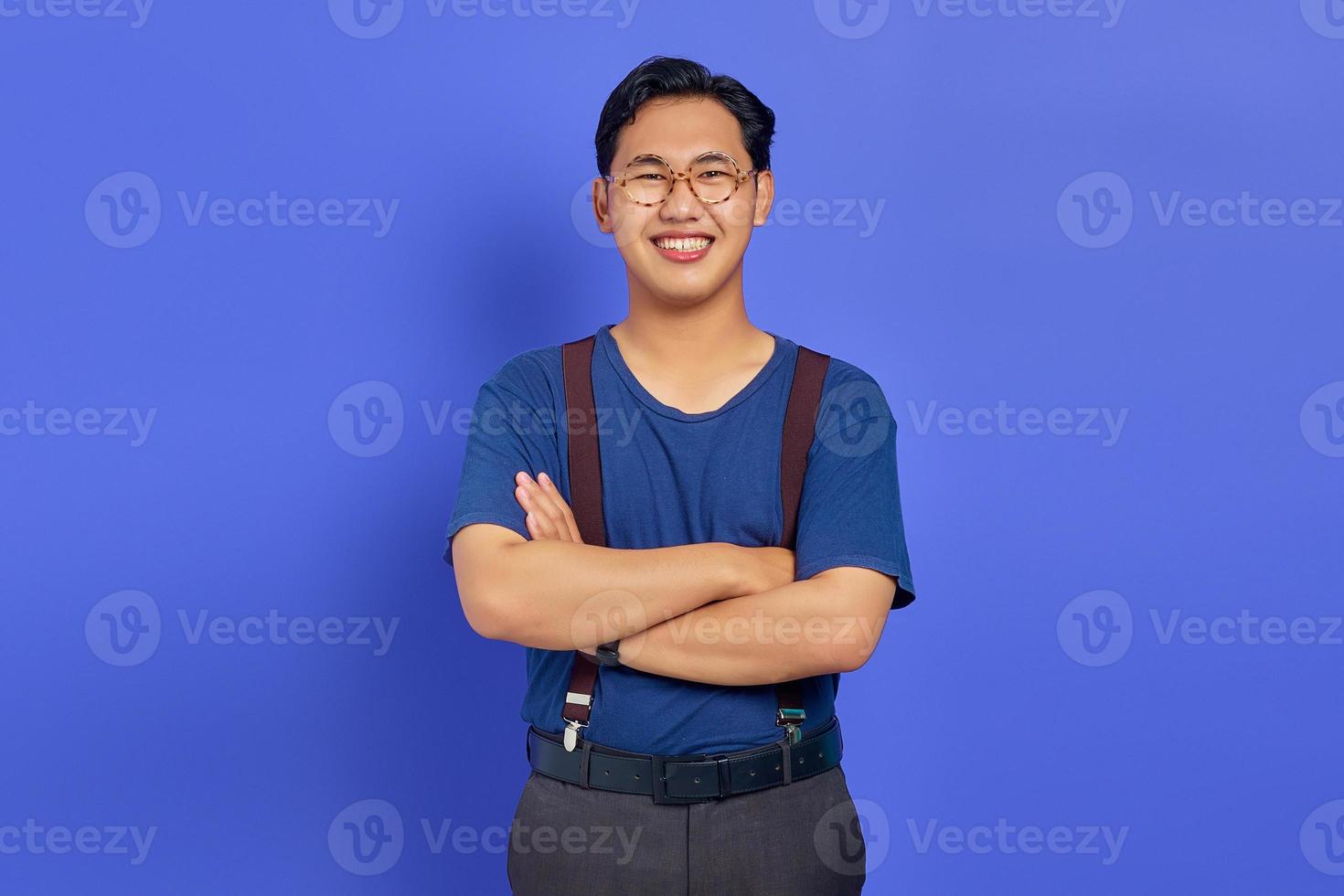 Porträt eines lächelnden jungen asiatischen Mannes, der seine Arme verschränkt und eine Brille auf violettem Hintergrund trägt foto