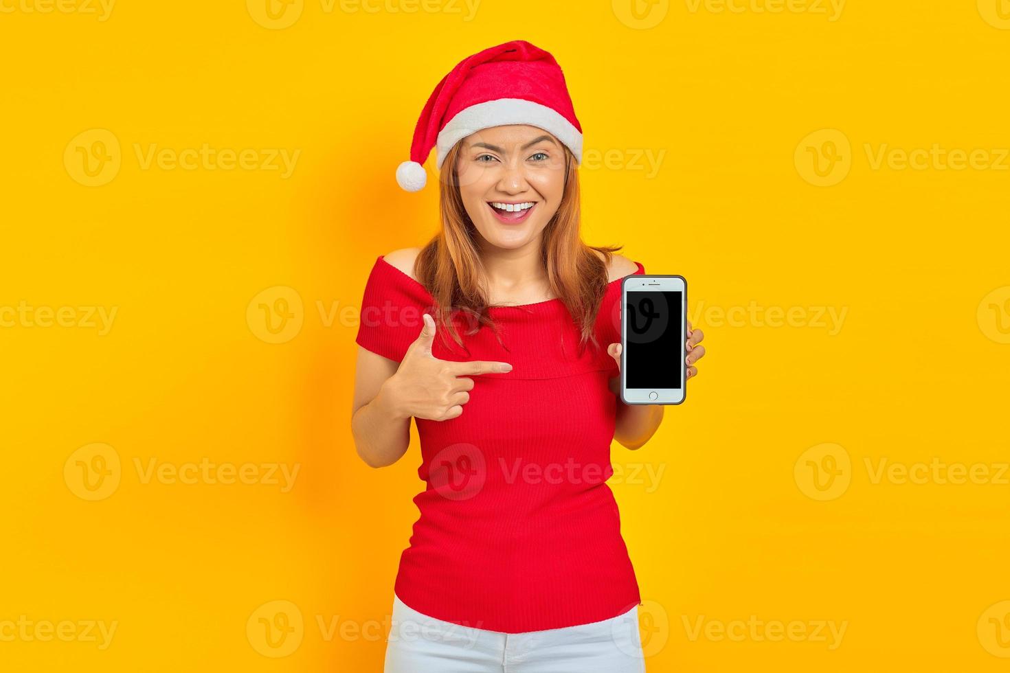 fröhliche junge asiatische frau in weihnachtsmannmütze, die mit dem finger auf ein leeres handy auf gelbem hintergrund zeigt foto