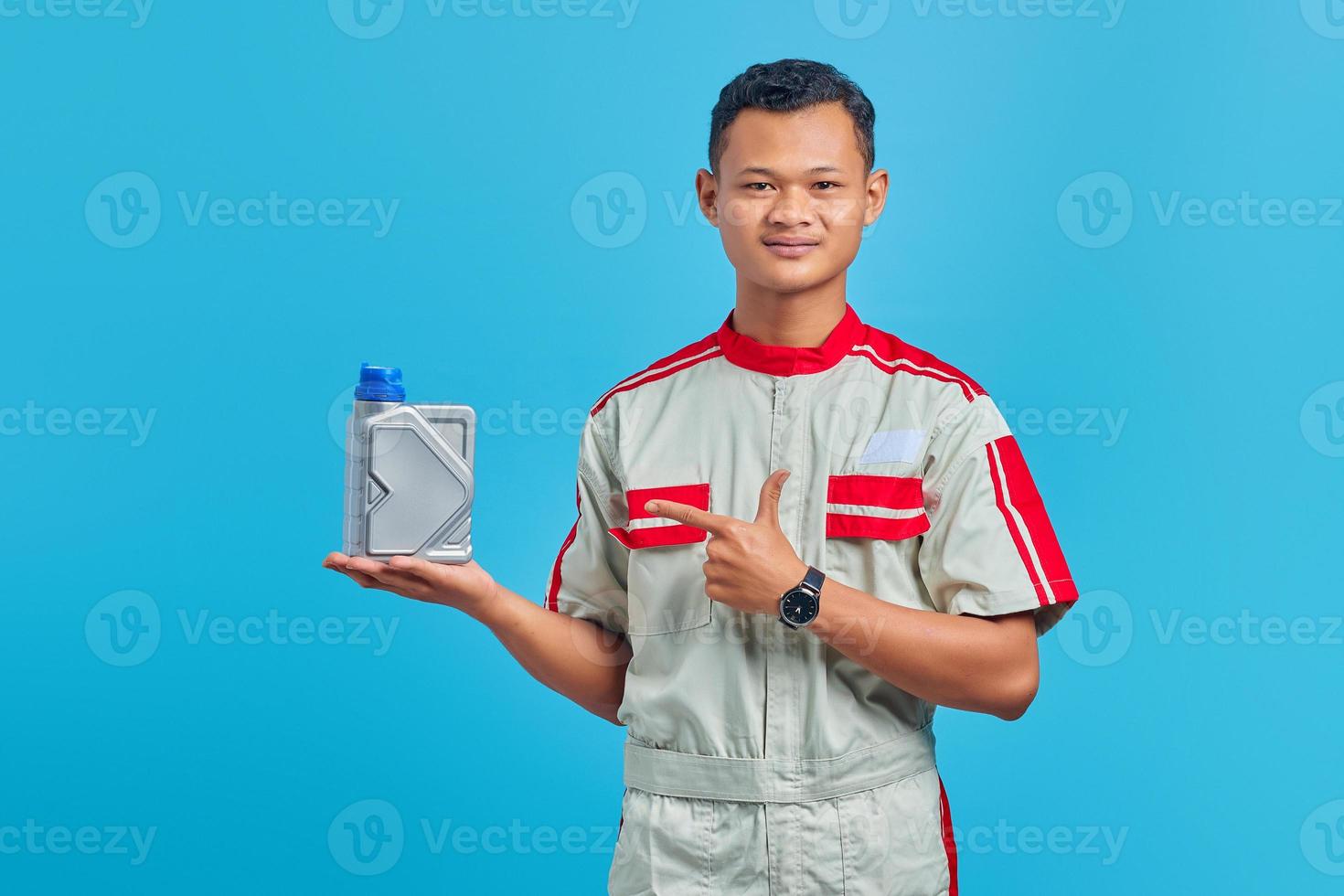 Porträt eines lächelnden jungen asiatischen Mechanikers, der mit dem Finger auf eine Plastikflasche Motoröl auf blauem Hintergrund zeigt foto