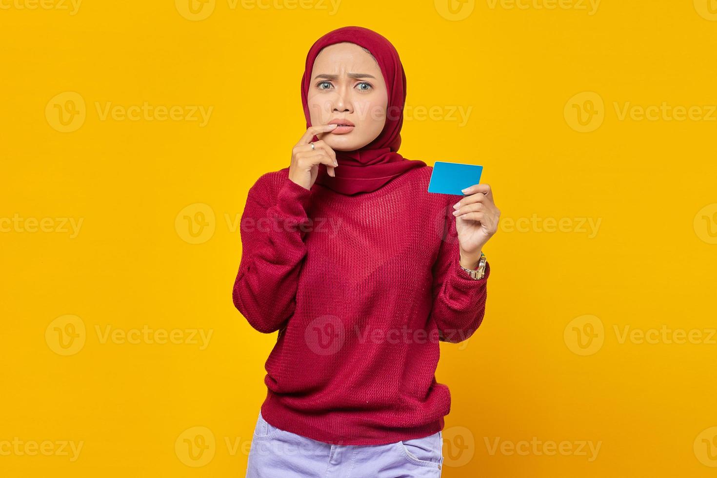 Schöne asiatische Frau, die gestresst und nervös aussieht, Nägel beißt und Kreditkarte auf gelbem Hintergrund zeigt foto