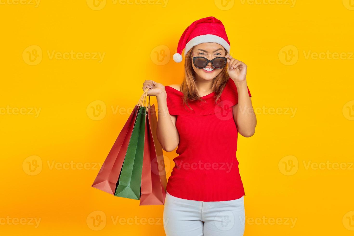 fröhliche junge asiatische frau in weihnachtsmannmütze und sonnenbrille mit einkaufstaschen einzeln auf gelbem hintergrund foto