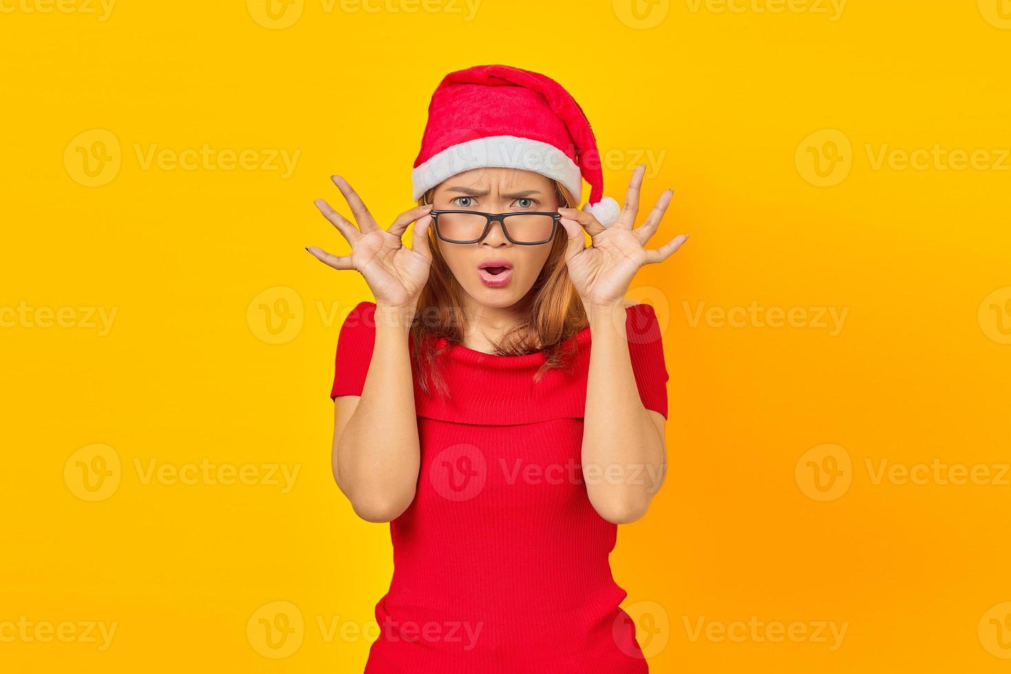 überraschte junge asiatische frau mit weihnachtsmütze mit brille bei offenem mund auf gelbem hintergrund foto