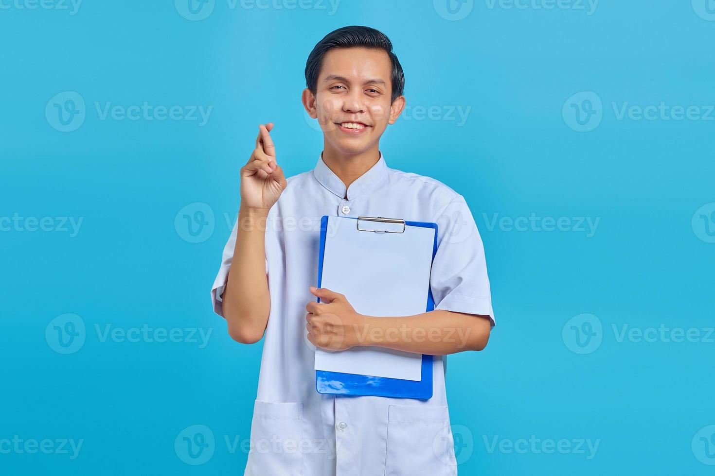 Porträt eines glücklichen jungen asiatischen Krankenpflegers, der die Finger kreuzt und die Zwischenablage auf blauem Hintergrund zeigt foto