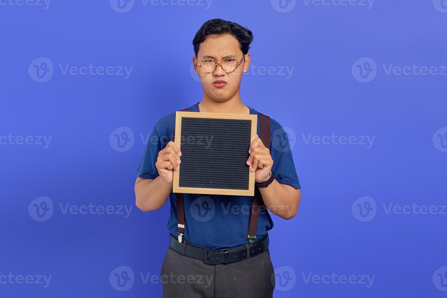 verwirrter hübscher junger Mann, der leeres Brett auf violettem Hintergrund zeigt foto