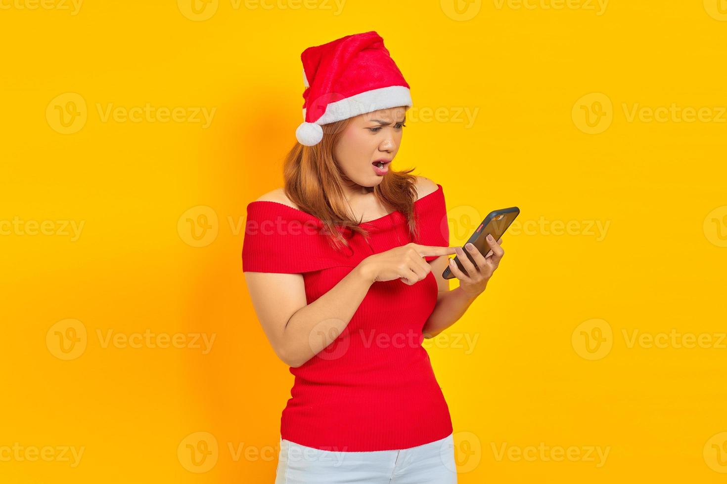 überraschte junge asiatische frau in weihnachtsmannmütze, die mit dem finger auf das handy zeigt, das über gelbem hintergrund isoliert ist foto