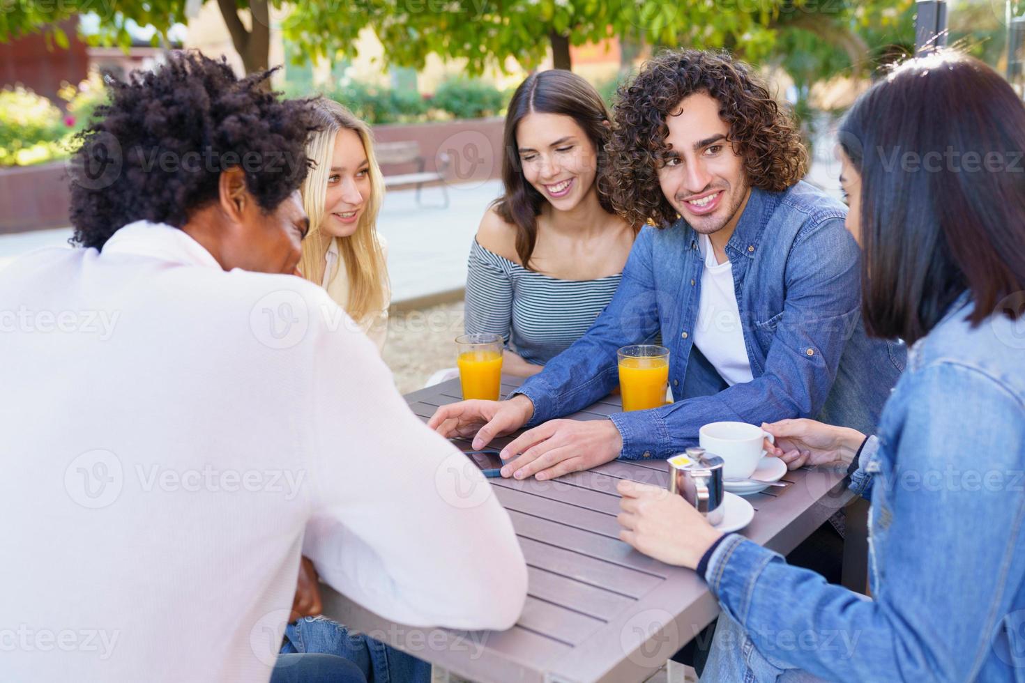 Typ zeigt seiner Gruppe von Freunden sein Smartphone, während er in einer Bar im Freien etwas trinkt foto