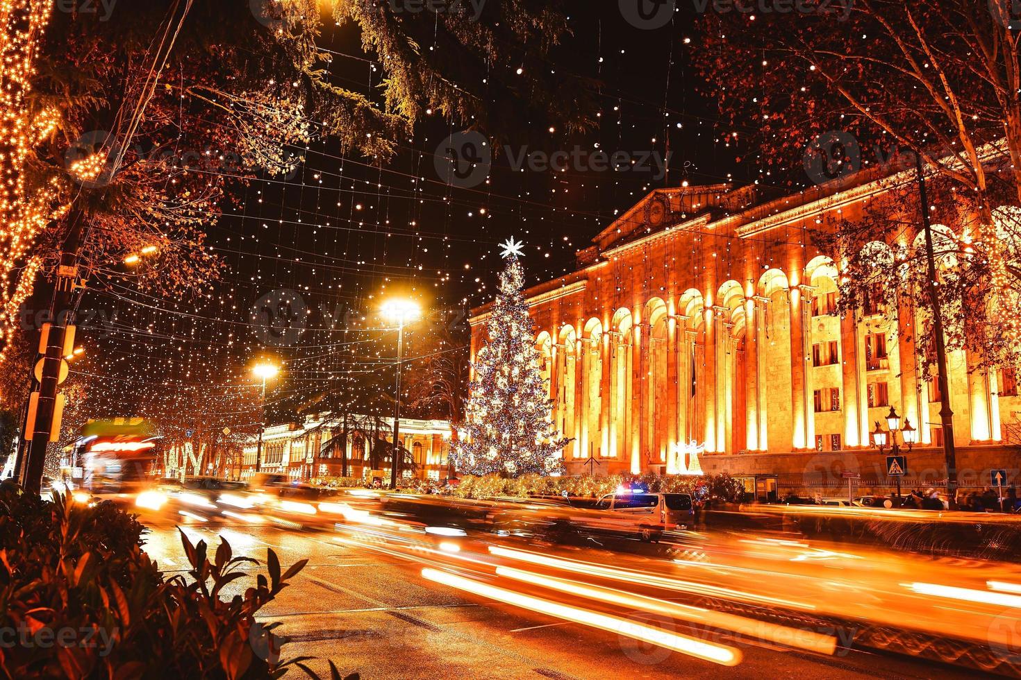 Autos im Verkehr zu Hauptverkehrszeiten in der Nacht in der Rustaveli Avenue in der Hauptstadt Tiflis in Georgien an Weihnachten mit Weihnachtsbaum durch das Parlament im Hintergrund foto