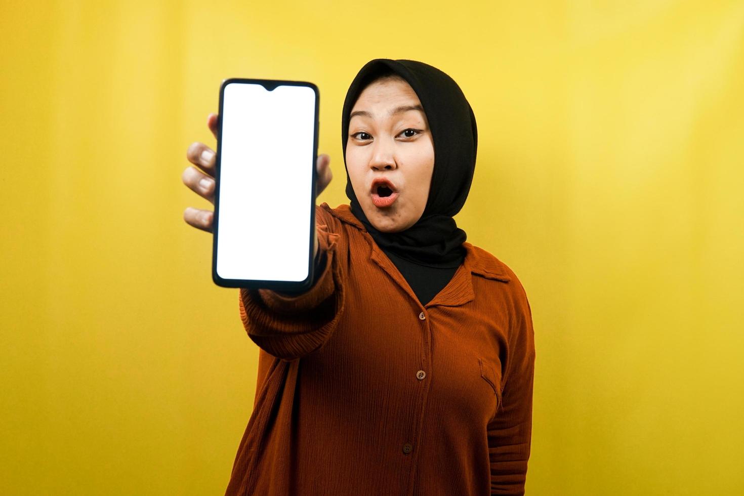 schöne junge asiatische muslimische frau schockiert, überrascht, wow-ausdruck, hand hält smartphone mit weißem oder leerem bildschirm, fördert app, fördert produkt, präsentiert etwas, isoliert foto