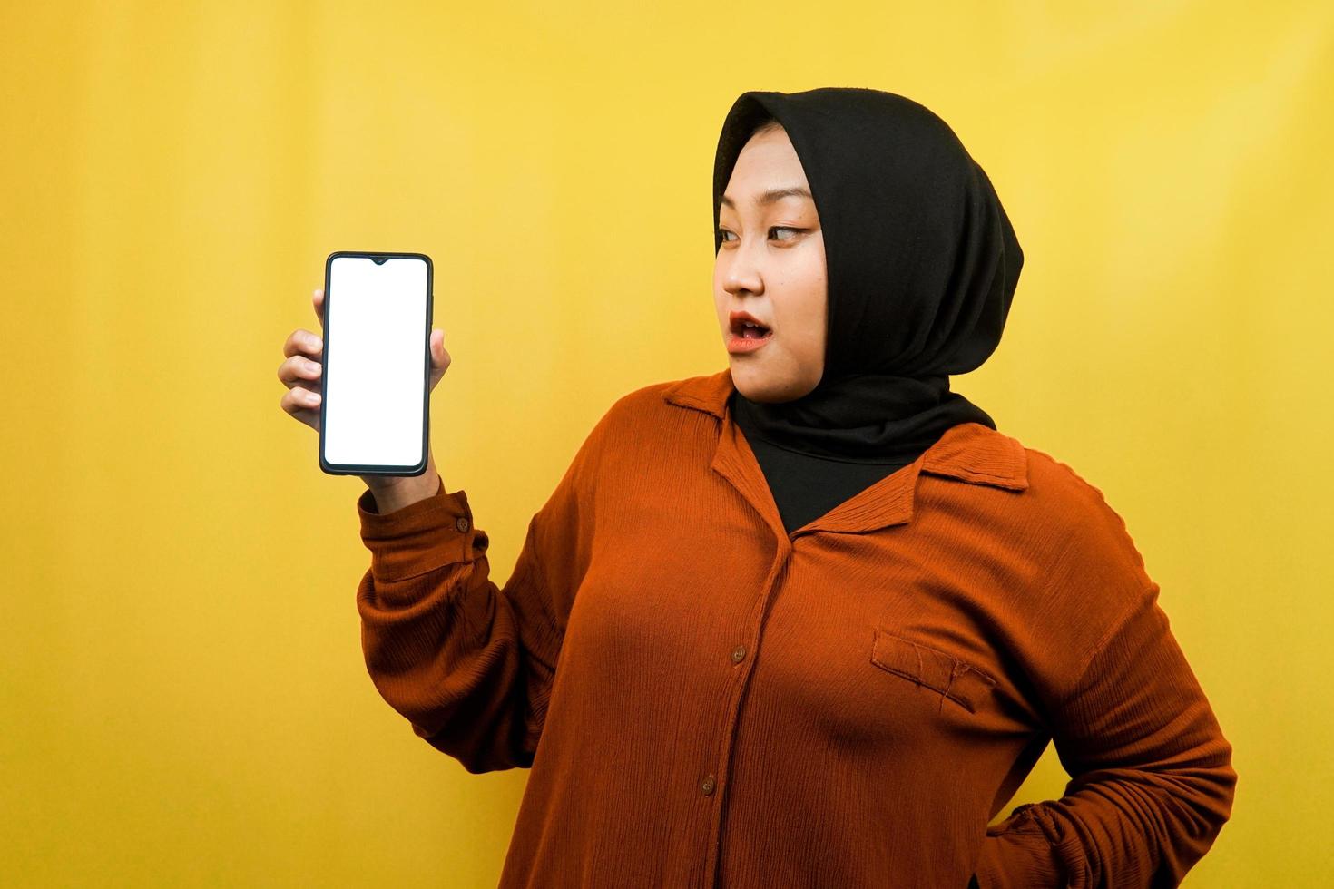 schöne junge asiatische muslimische frau schockiert, überrascht, wow-ausdruck, hand hält smartphone mit weißem oder leerem bildschirm, fördert app, fördert produkt, präsentiert etwas, isoliert foto