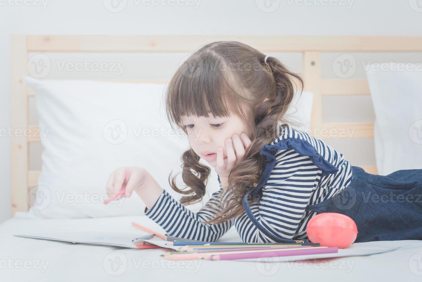 süßes kleines Mädchen, das Hausaufgaben macht, mit bunten Bleistiften auf dem Bett zu Hause schreibt und zeichnet. elegantes Design für spielende Kinder, Vorschule lernen zu schreiben, zu lesen und kreatives Kunsterziehungskonzept foto