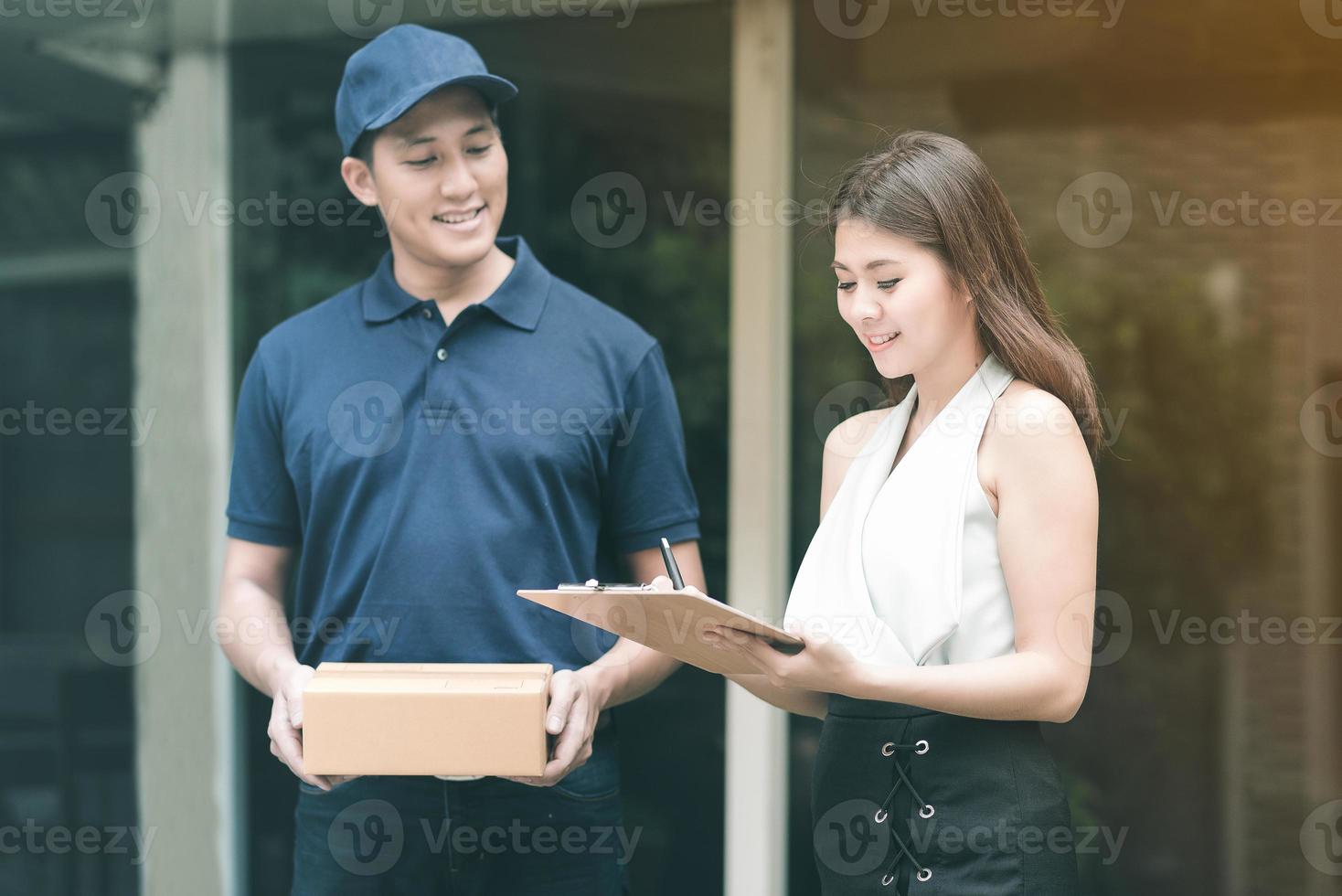 hübscher junger asiatischer liefermann, der lächelt und einen karton hält, während er wartet, schöne junge asiatische frau, die unterschrift in die zwischenablage setzt, um den empfang zu bestätigen. foto