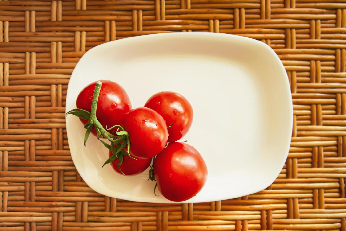 Eine Ansammlung von roten Kirschtomaten auf einem weißen flachen rechteckigen Teller steht auf einer hölzernen Korbfläche. Sicht von oben. Komposition des Lebensstils. foto