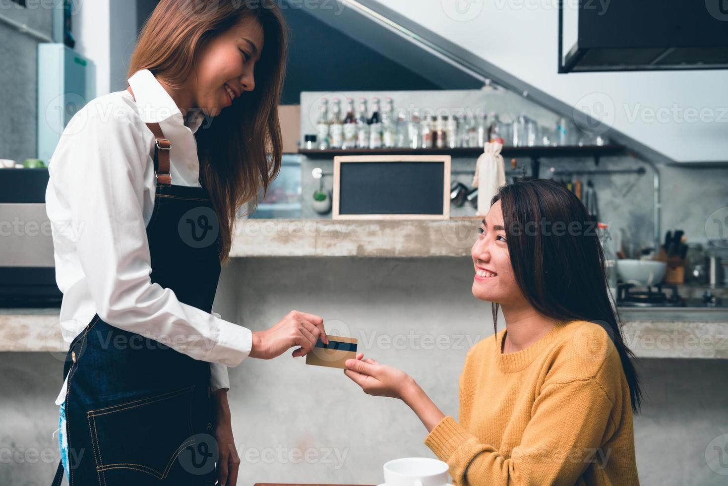 junge asiatische frau leistet eine zahlung an den cafébesitzer per kreditkarte mit dem hintergrund einer getränketheke in einem kleinen café. junge frau bezahlt ihren kaffee mit kreditkarte. Essen und Trinken. foto