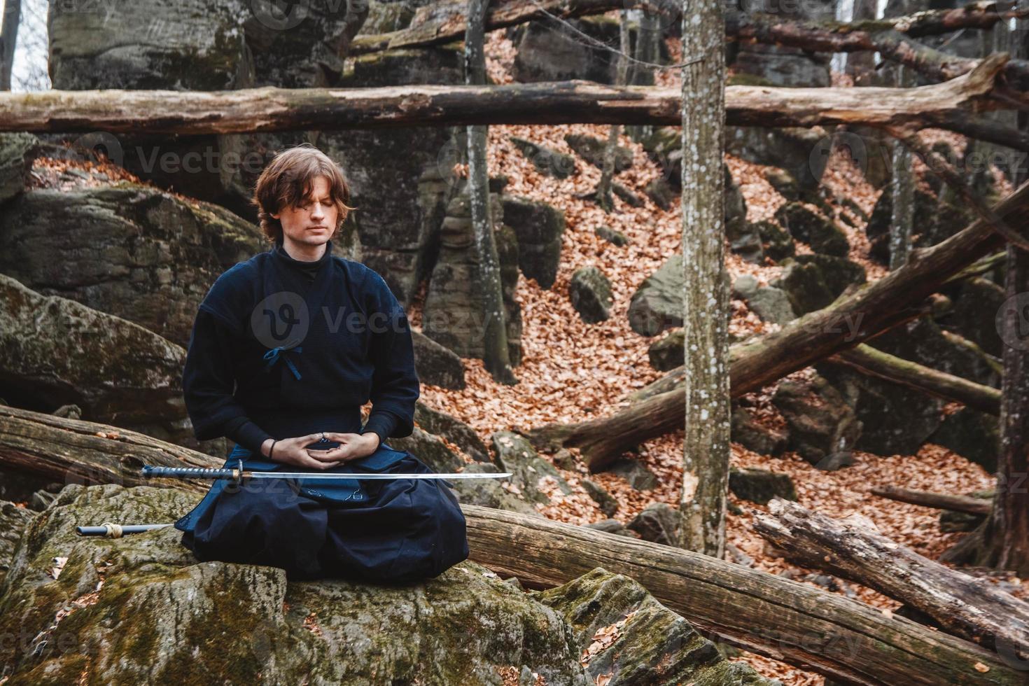 Mann im schwarzen Kimono mit Schwert meditiert und konzentriert sich auf Felsen und Waldhintergrund foto