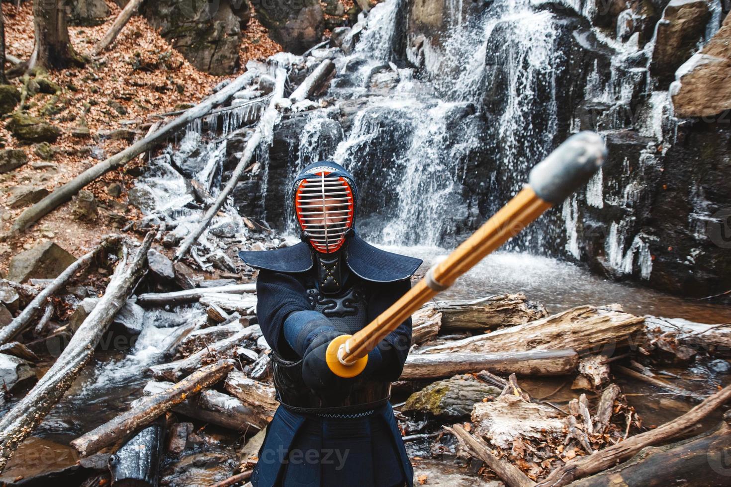 Mann, der Kendo mit Bambusschwert auf Wasserfall, Felsen und Waldhintergrund übt foto