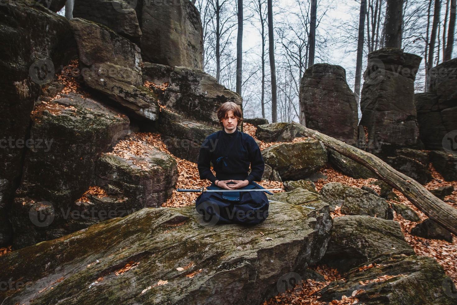 Mann im schwarzen Kimono mit Schwert meditiert und konzentriert sich auf Felsen und Waldhintergrund foto