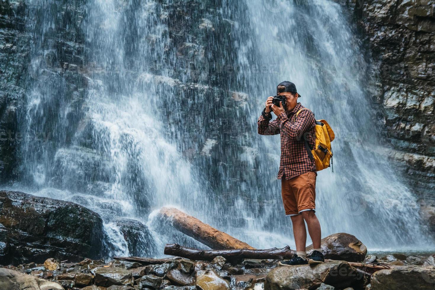 Mann mit gelbem Rucksack, der auf dem Hintergrund eines Wasserfalls steht, macht eine Fotolandschaft foto