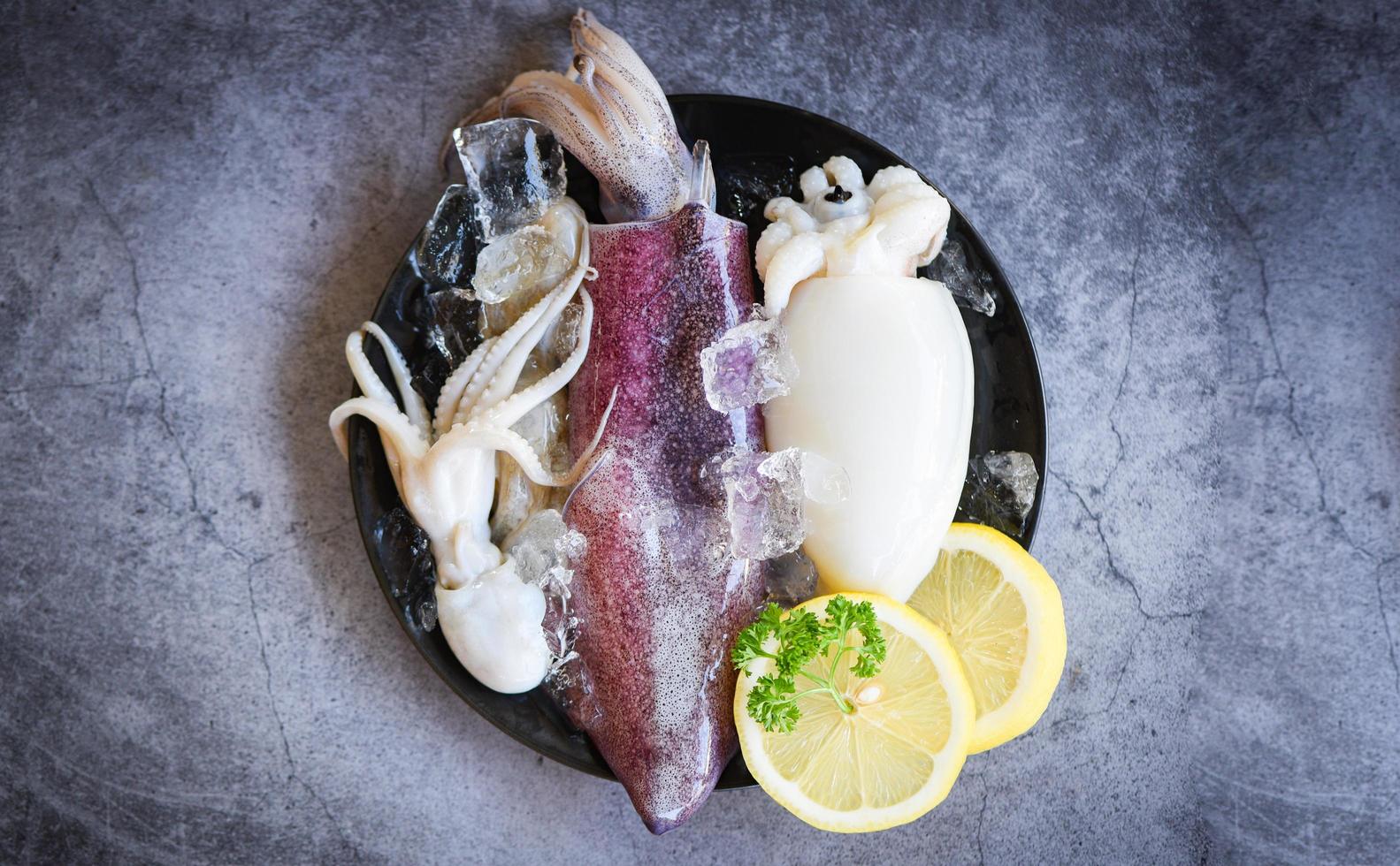 frischer Tintenfisch, Tintenfisch oder Tintenfisch für gekochtes Essen im Restaurant oder auf dem Fischmarkt - roher Tintenfisch mit Eis und Salatgewürzen Zitrone auf dem schwarzen Plattenhintergrund foto