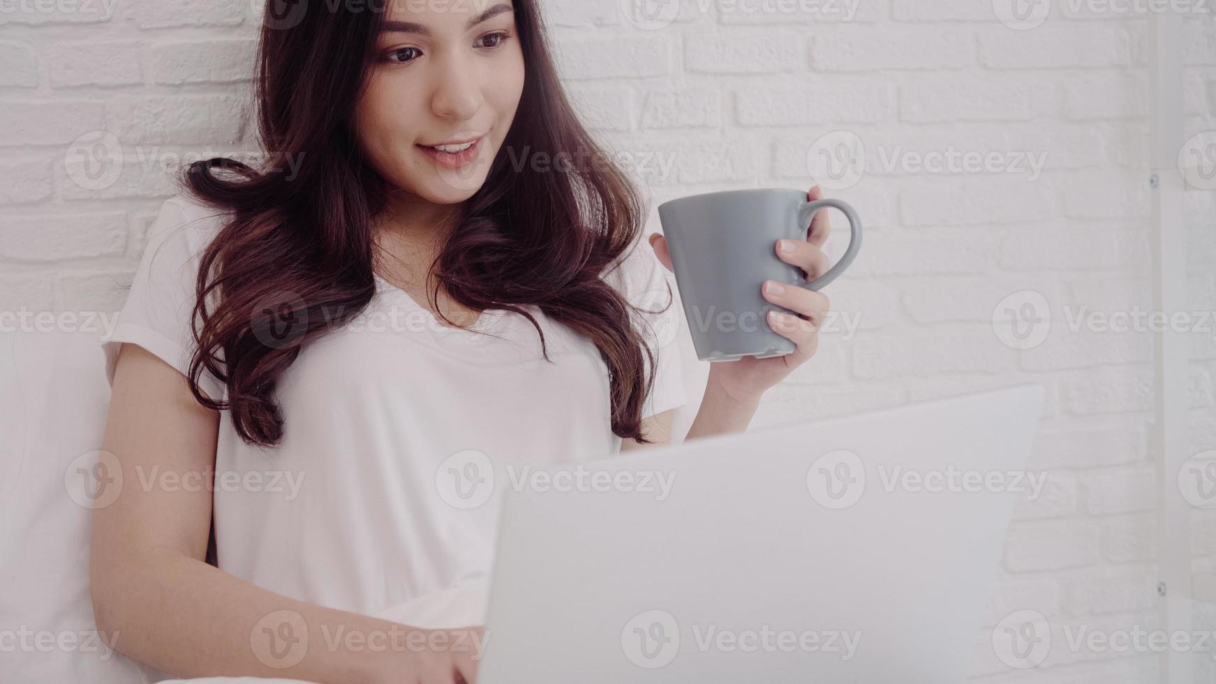 Porträt einer schönen attraktiven asiatischen Frau mit Computer oder Laptop, die eine warme Tasse Kaffee oder Tee hält, während sie auf dem Bett liegt, wenn sie sich zu Hause in ihrem Schlafzimmer entspannt. Lifestyle-Frauen zu Hause Konzept. foto