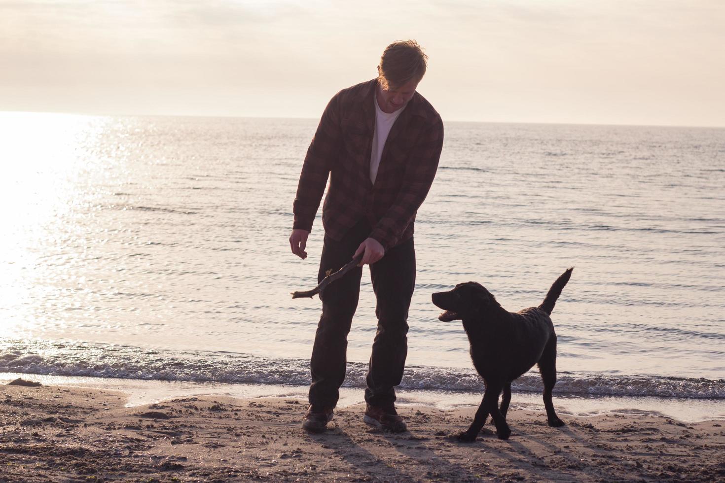 junger Mann, der mit schwarzem Hund am Morgenstrand spazieren geht foto