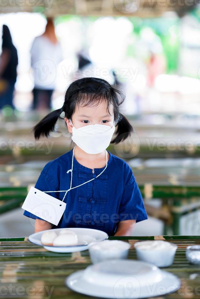 Kinder mit weißer Gesichtsmaske sitzen und warten auf den Unterricht zur Lebensmittelkonservierung im Unterricht. Kind schwitzt, nasse Haarsträhnen auf der Stirn. vertikales Bild. foto
