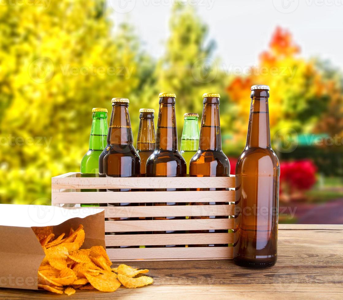 Bierflaschen und Kartoffelchips auf Holztisch mit verschwommenem Wald im Hintergrund, farbige Flasche, Speisen- und Getränkekonzept, selektiver Fokus, Kopierraum foto
