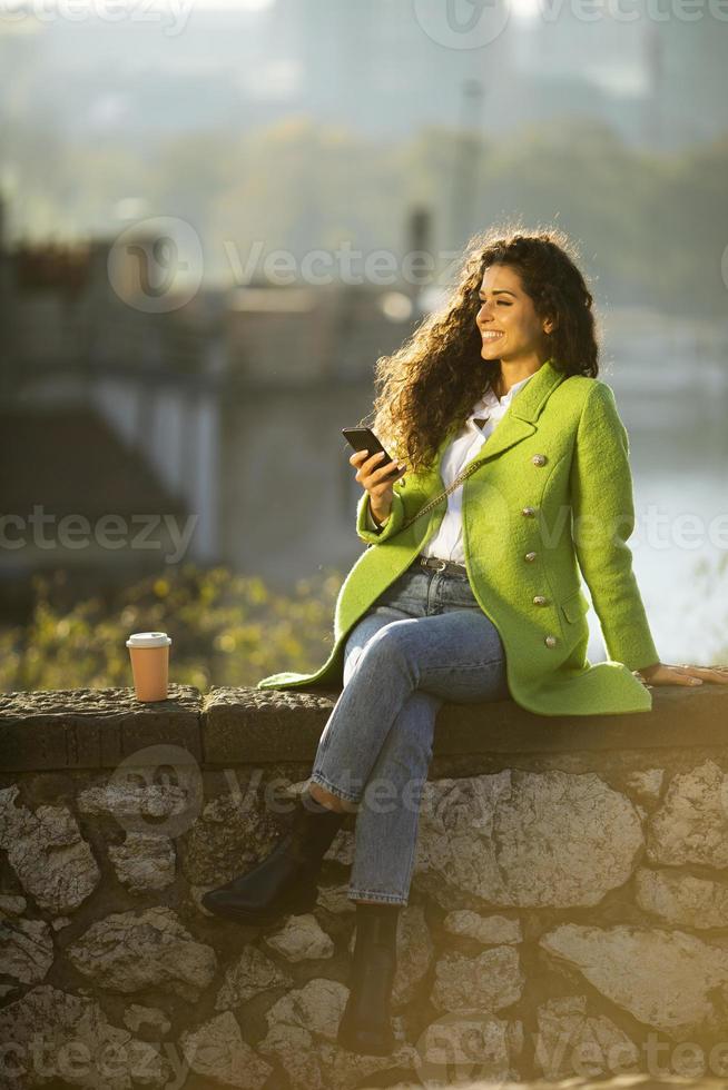 hübsche junge Frau mit Smartphone, während sie am Fluss sitzt und Kaffee zum Mitnehmen trinkt foto