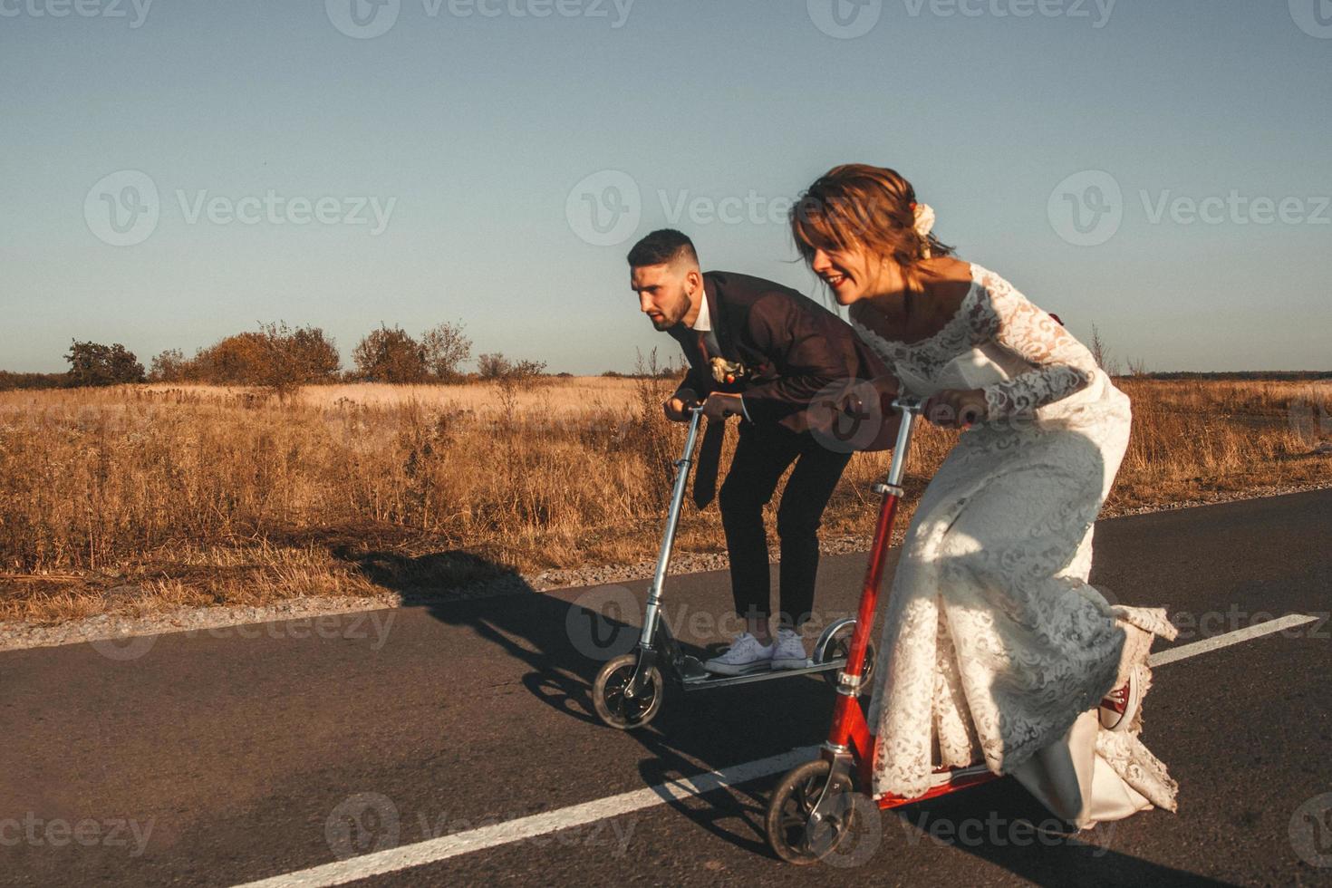 lächelndes Hochzeitspaar, das bei Sonnenuntergang auf Rollern entlang der Straße außerhalb der Stadt fährt. Platz für Text oder Werbung foto