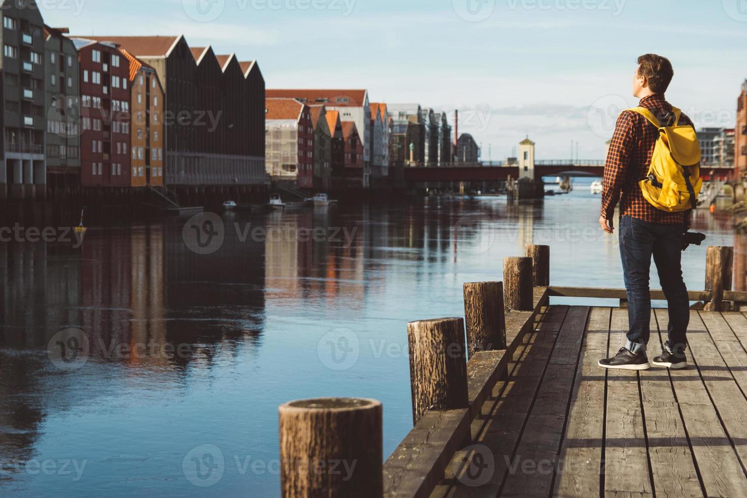 Fotograf mit gelbem Rucksack, der auf einem Holzsteg steht und ein Foto vor Stadt- und Flusshintergrund macht