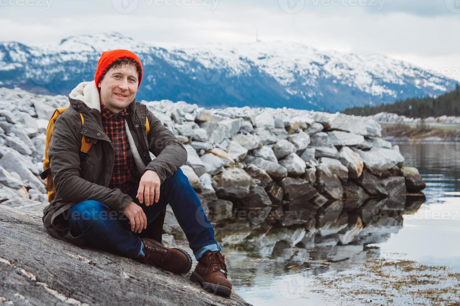 junger Mann mit gelbem Rucksack, der einen roten Hut trägt und am Ufer vor dem Hintergrund von Berg und See sitzt. Platz für Ihre Textnachricht oder Werbeinhalte. Reise-Lifestyle-Konzept. foto