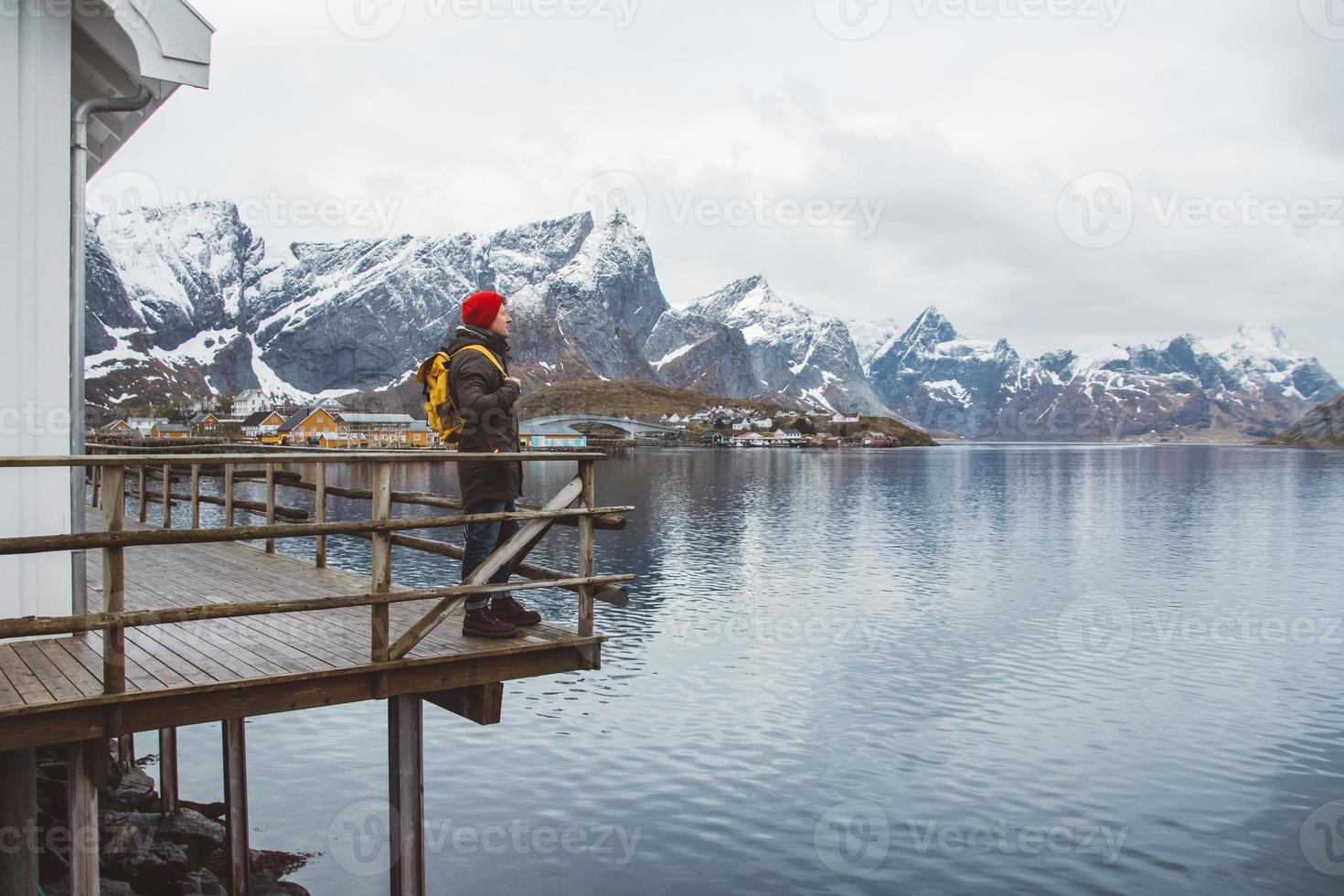 junger Mann mit einem Rucksack, der auf einem hölzernen Pier steht, der Hintergrund der schneebedeckten Berge und des Sees. Platz für Text oder Werbung foto