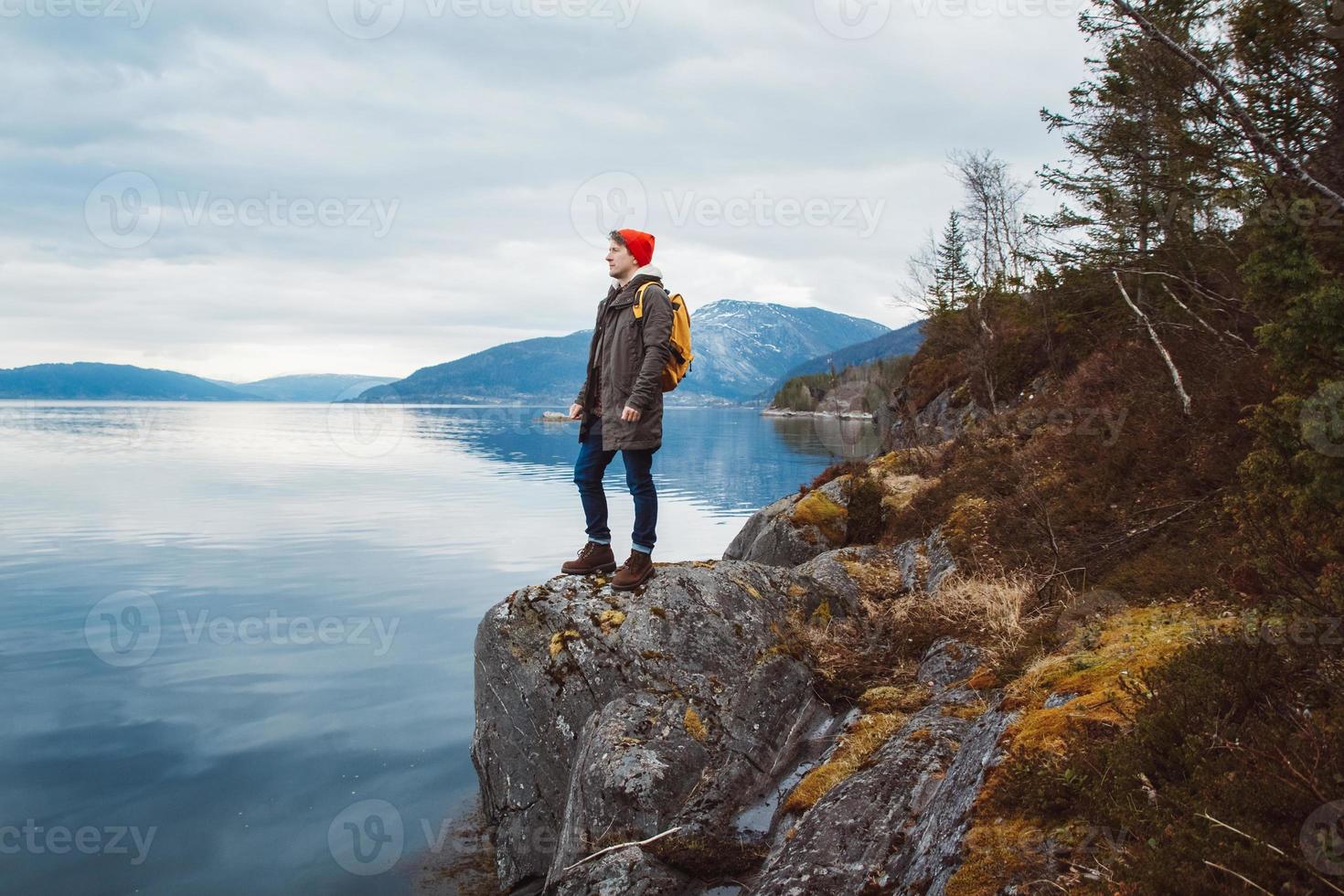 junger Mann mit gelbem Rucksack, der einen roten Hut trägt, der auf einem Felsen vor dem Hintergrund von Berg und See steht. Platz für Ihre Textnachricht oder Werbeinhalte. Reise-Lifestyle-Konzept foto