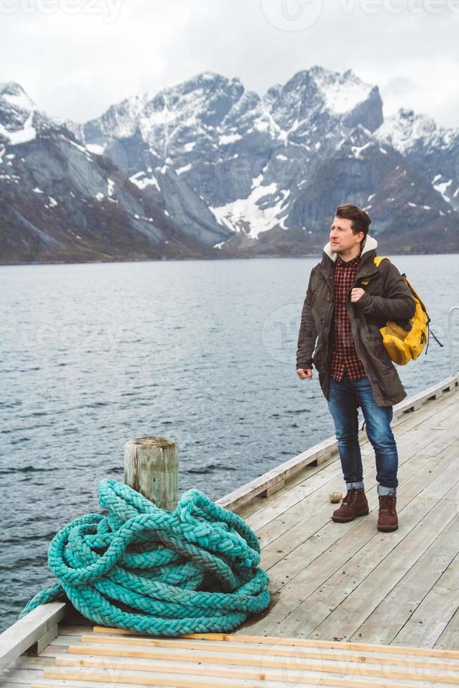 Reisender Mann mit einem Rucksack, der auf einem Holzsteg steht, vor dem Hintergrund der schneebedeckten Berge und des Sees. Platz für Text oder Werbung foto