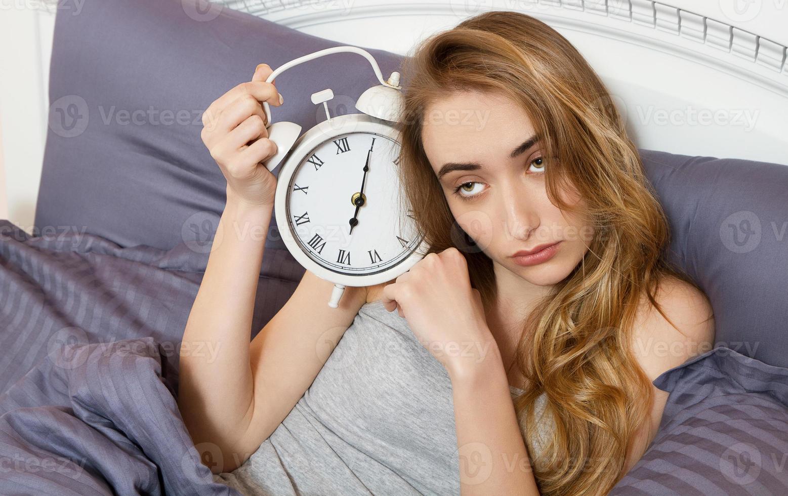 junges schönes verschlafenes mädchen, das eine uhr hält und morgens in ihrem bett im schlafzimmer liegt. foto