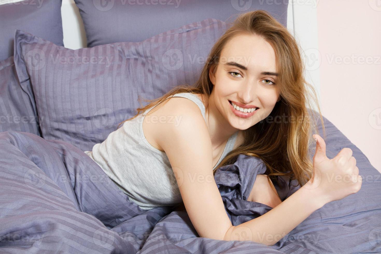 Porträt eines schönen Lächelnmädchens auf dem Bett nach dem Schlafen, Frau zeigt wie Schild im Bett foto