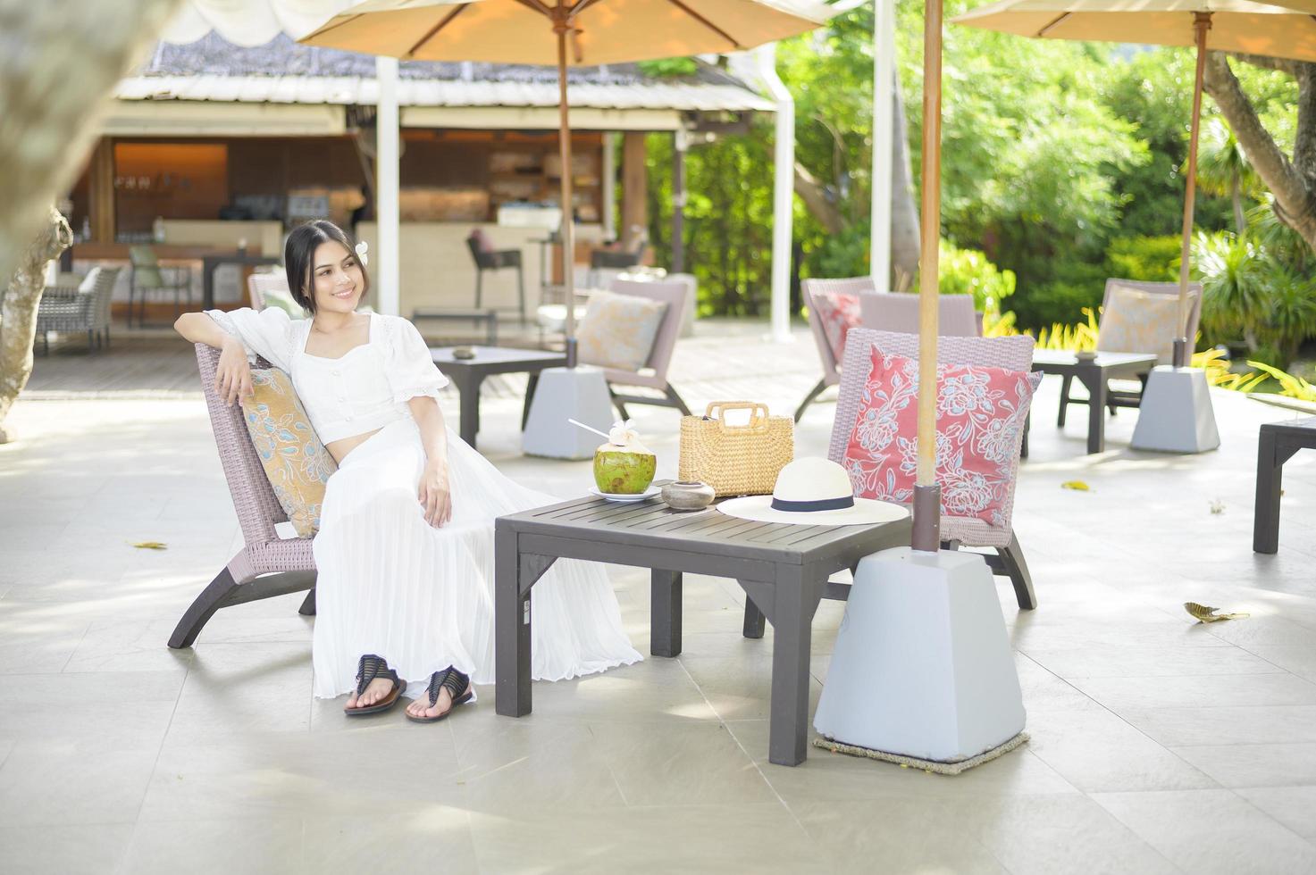 schöne Touristin mit weißer Blume auf ihrem Haar, die während der Sommerferien Kokosnuss trinkt, die auf einem Liegestuhl sitzt foto