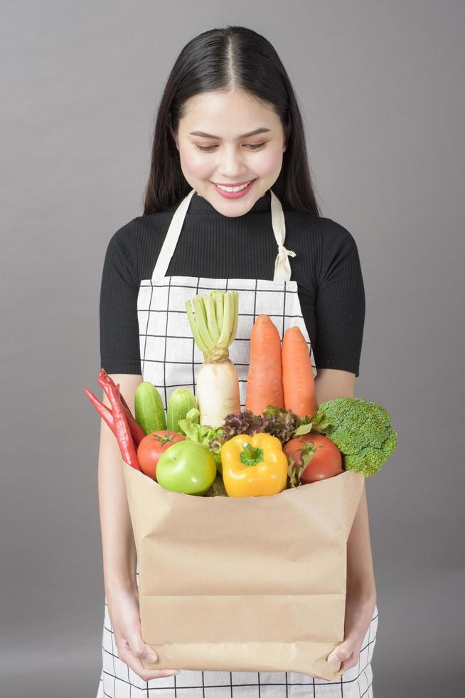 Porträt der schönen jungen Frau mit Gemüse in der Einkaufstüte im grauen Studiohintergrund foto