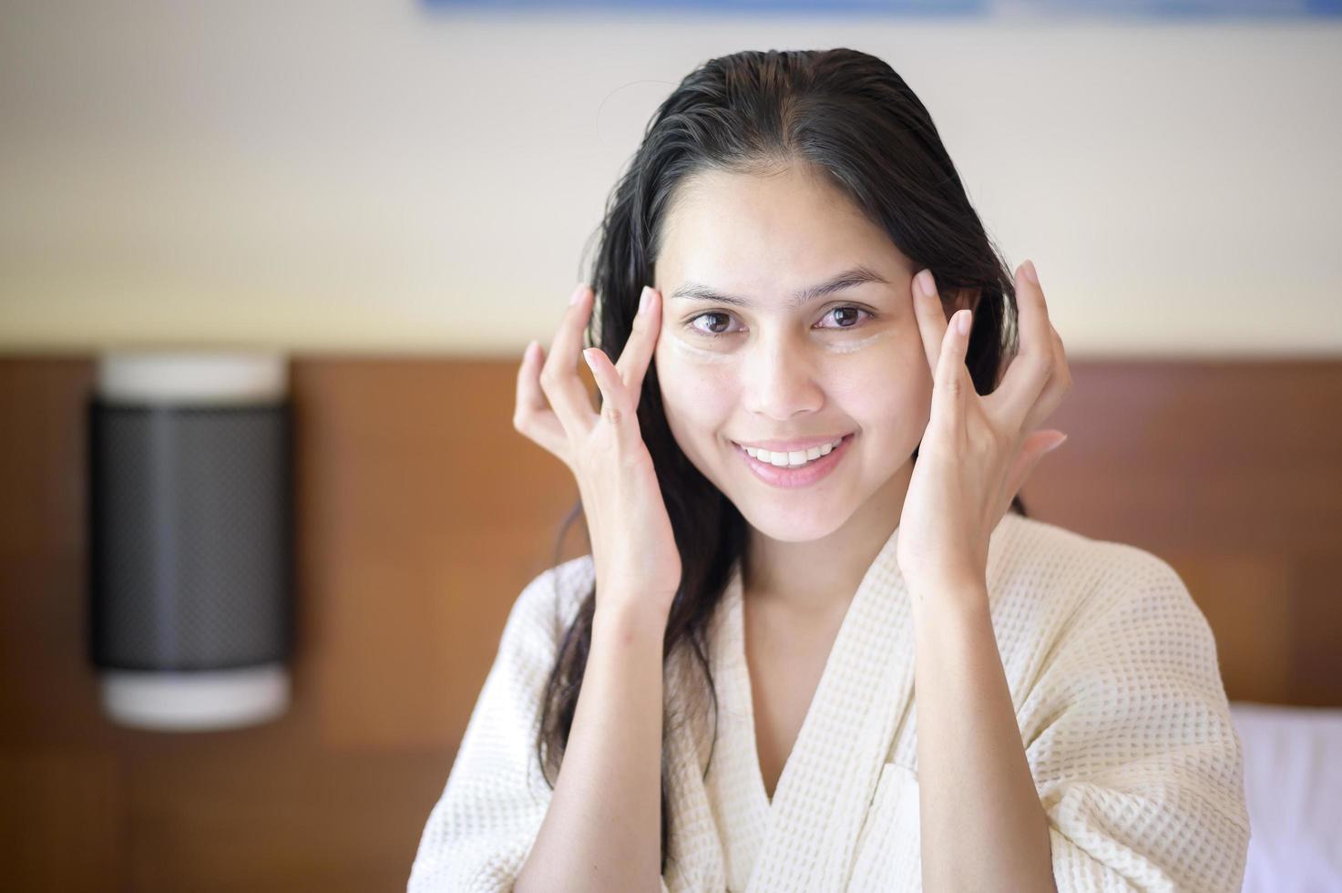 Eine glückliche schöne Frau im weißen Bademantel, die Feuchtigkeitscreme auf das Gesicht im Schlafzimmer, Hautpflege- und Behandlungskonzept aufträgt foto