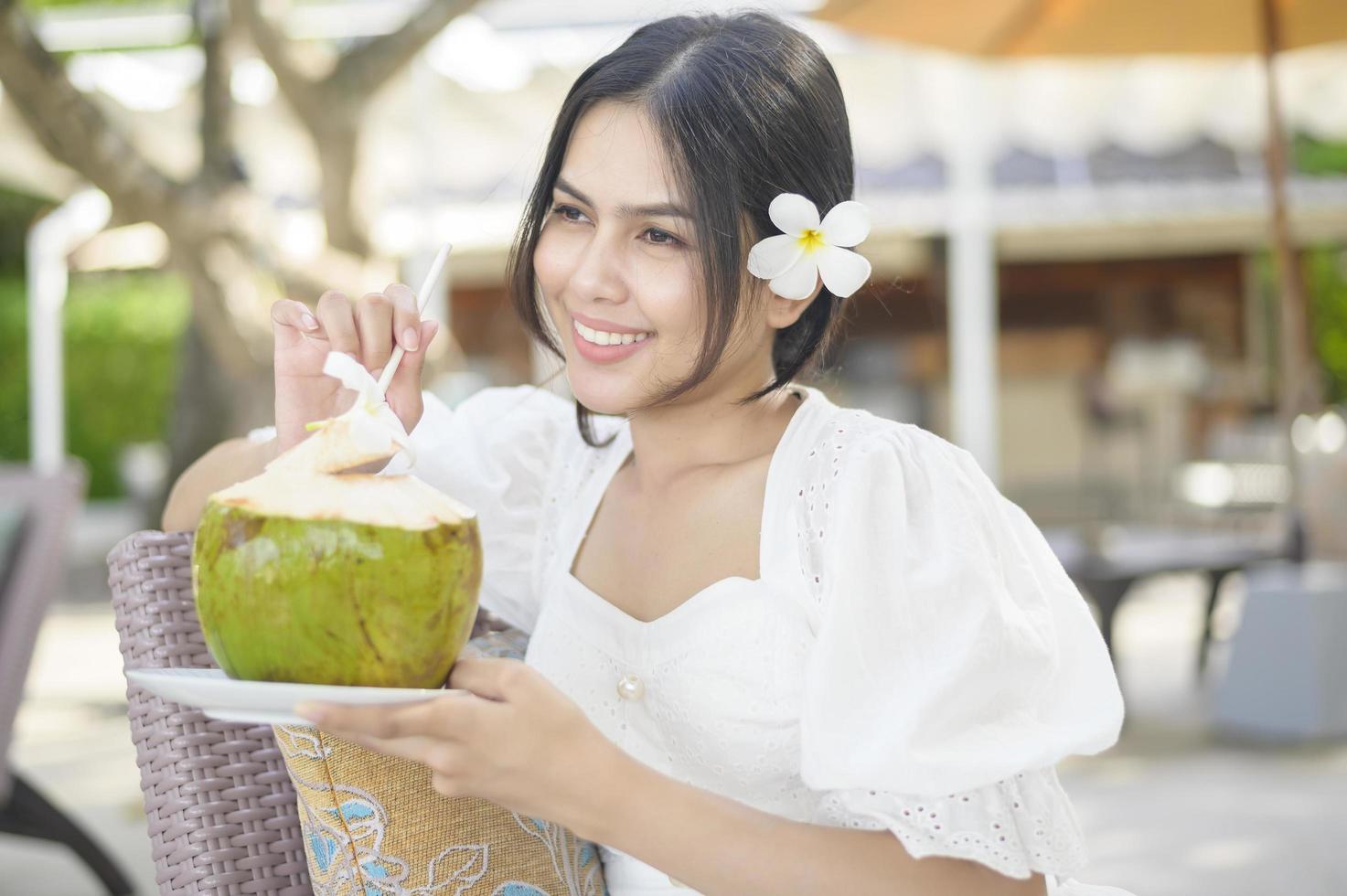 schöne Touristin mit weißer Blume auf ihrem Haar, die während der Sommerferien Kokosnuss trinkt, die auf einem Liegestuhl sitzt foto