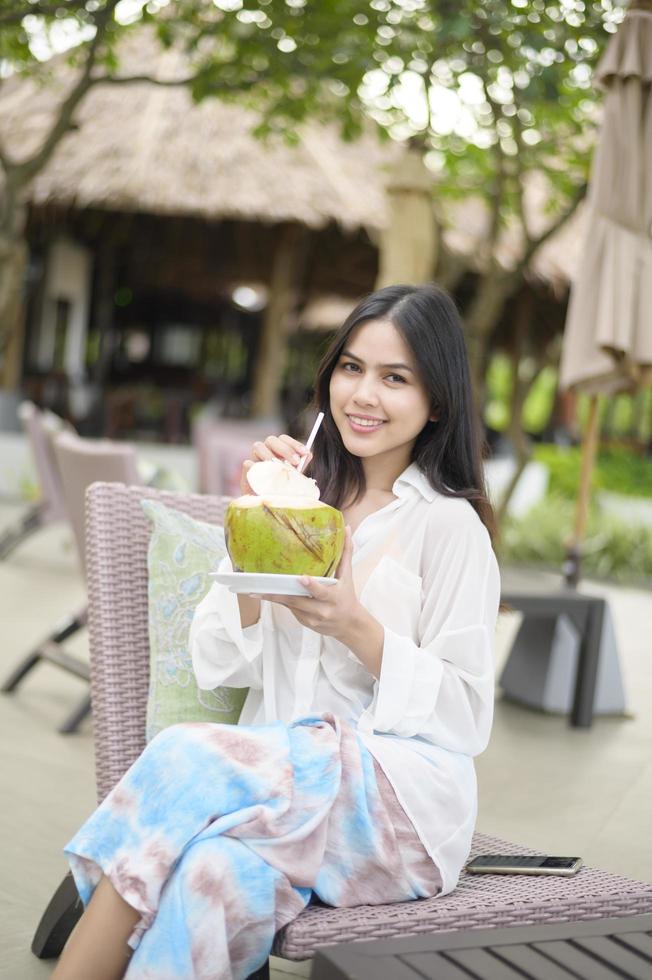 schöne Touristin mit weißer Blume auf ihrem Haar, die während der Sommerferien Kokosnuss trinkt, die auf einem Liegestuhl sitzt foto