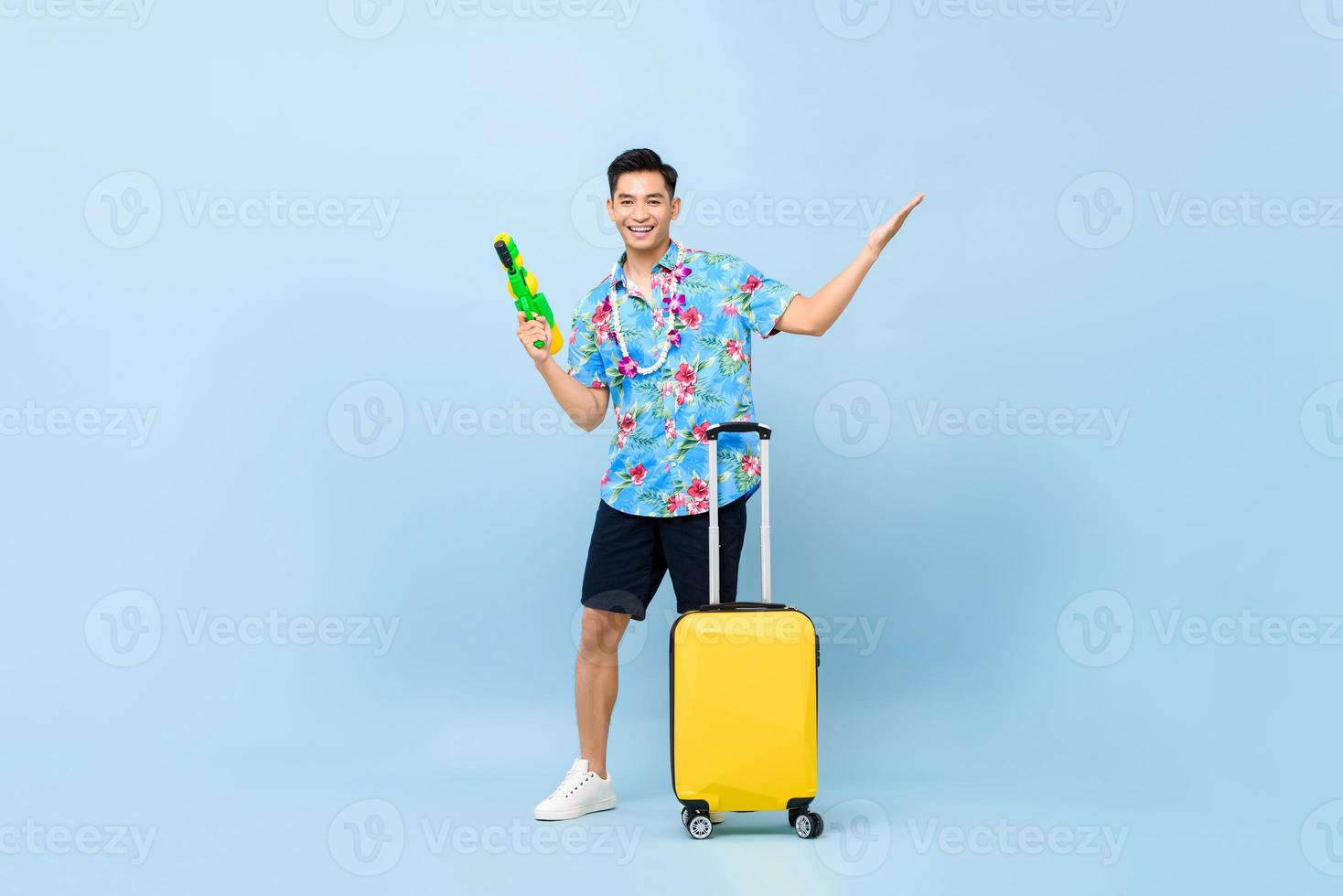 lächelnder hübscher asiatischer Touristenmann, der mit Wasserpistole und Gepäck während des Songkran-Festivals reist, schoss blauen Hintergrund foto