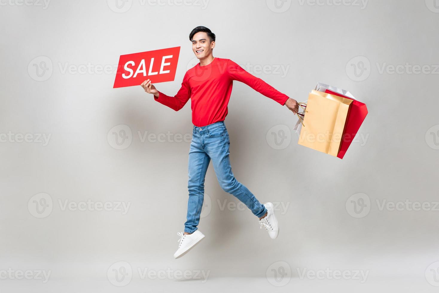 Überraschter asiatischer Mann, der Einkaufstüten und rotes Verkaufsschild hält, das in isoliertem hellgrauem Studiohintergrund für das chinesische Neujahrsverkaufskonzept springt foto