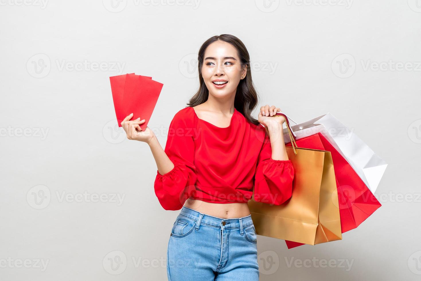 glückliche asiatische frau, die einkaufstaschen und rote umschläge einzeln in grauem studiohintergrund für chinesisches neues jahresverkaufskonzept hält foto