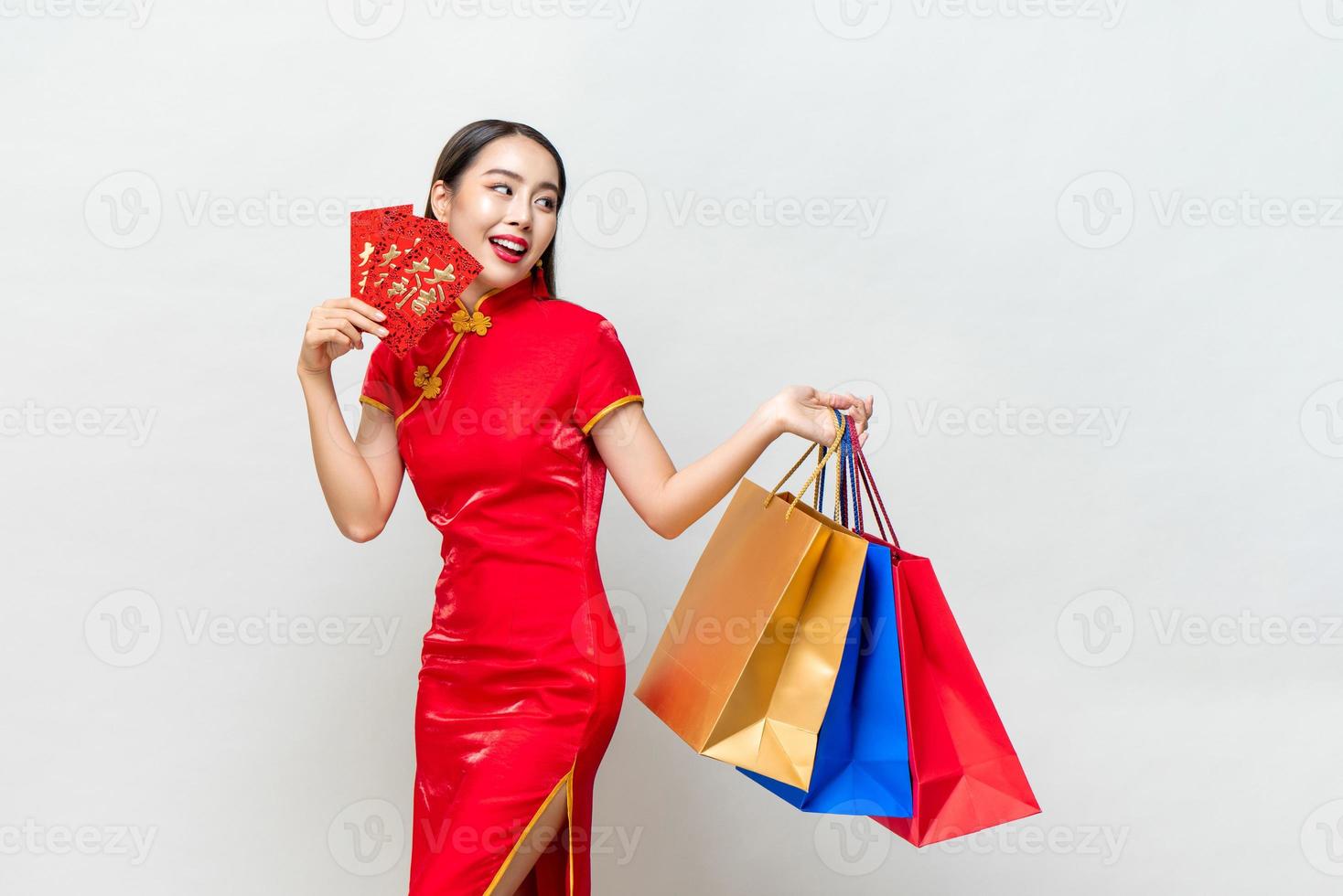 asiatische Frau in traditioneller Kleidung, die Taschen und rote Umschläge in isoliertem hellgrauem Studiohintergrund für das chinesische Neujahrs-Einkaufskonzept hält, Text bedeutet viel Glück, großen Gewinn foto