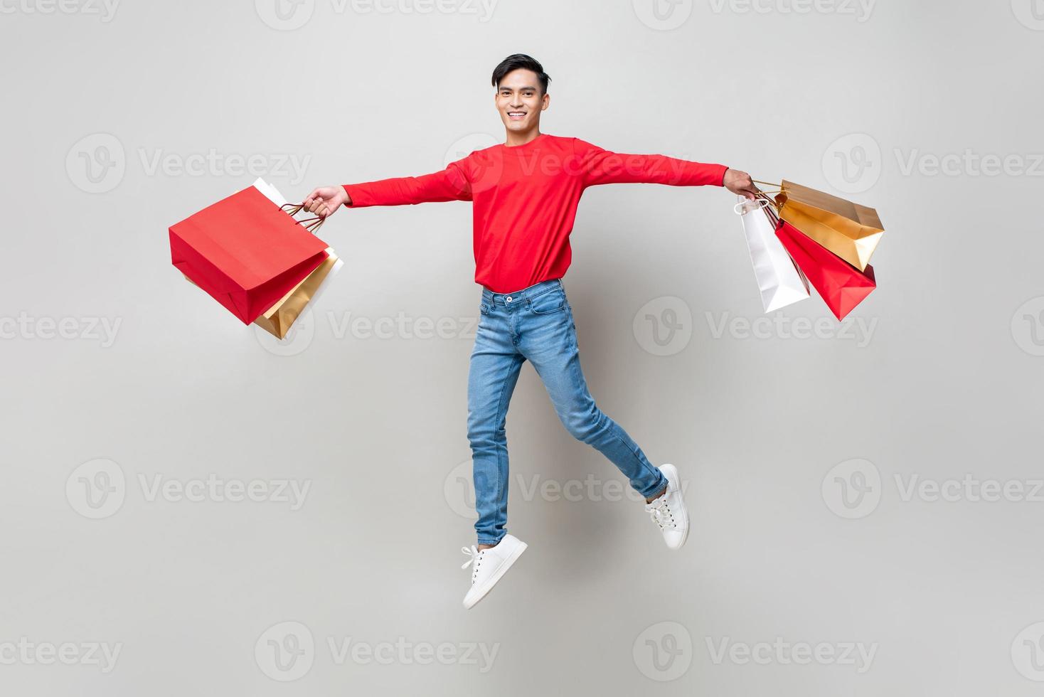 Schöner lächelnder asiatischer Mann, der Einkaufstaschen hält und in isoliertem Studio hellgrauen Hintergrund für chinesische Neujahrsverkaufskonzepte springt foto