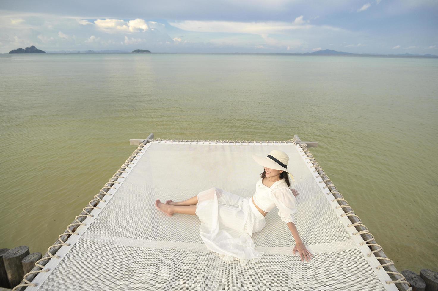 Eine glückliche, schöne Frau im weißen Kleid, die auf der Terrasse der tropischen Insel und dem türkisfarbenen Meer, Sommer- und Urlaubskonzept genießt und sich entspannt foto