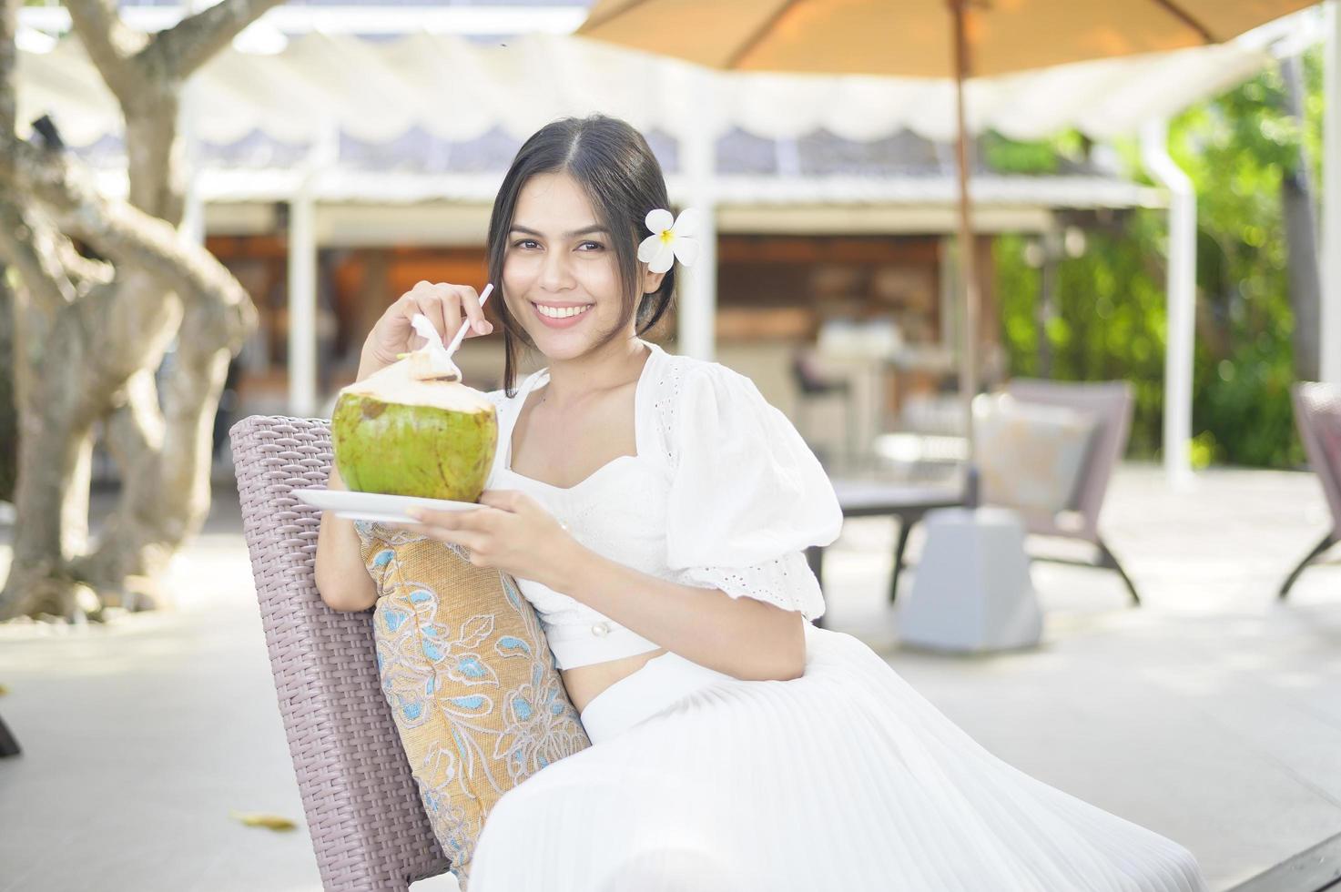 schöne Touristin mit weißer Blume auf ihrem Haar, die während der Sommerferien Kokosnuss trinkt, die auf einem Liegestuhl sitzt foto