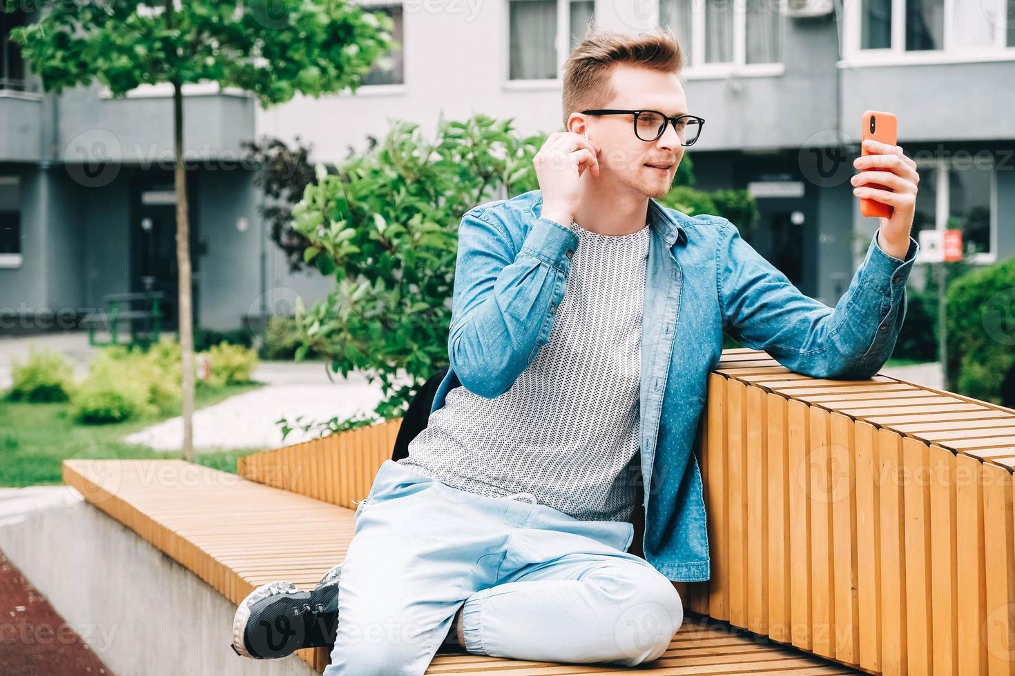 Mann in Hemd, Jeans und Brille, der auf einem Smartphone kommuniziert, das auf einer Bank vor einem Hintergrund von Häusern sitzt foto