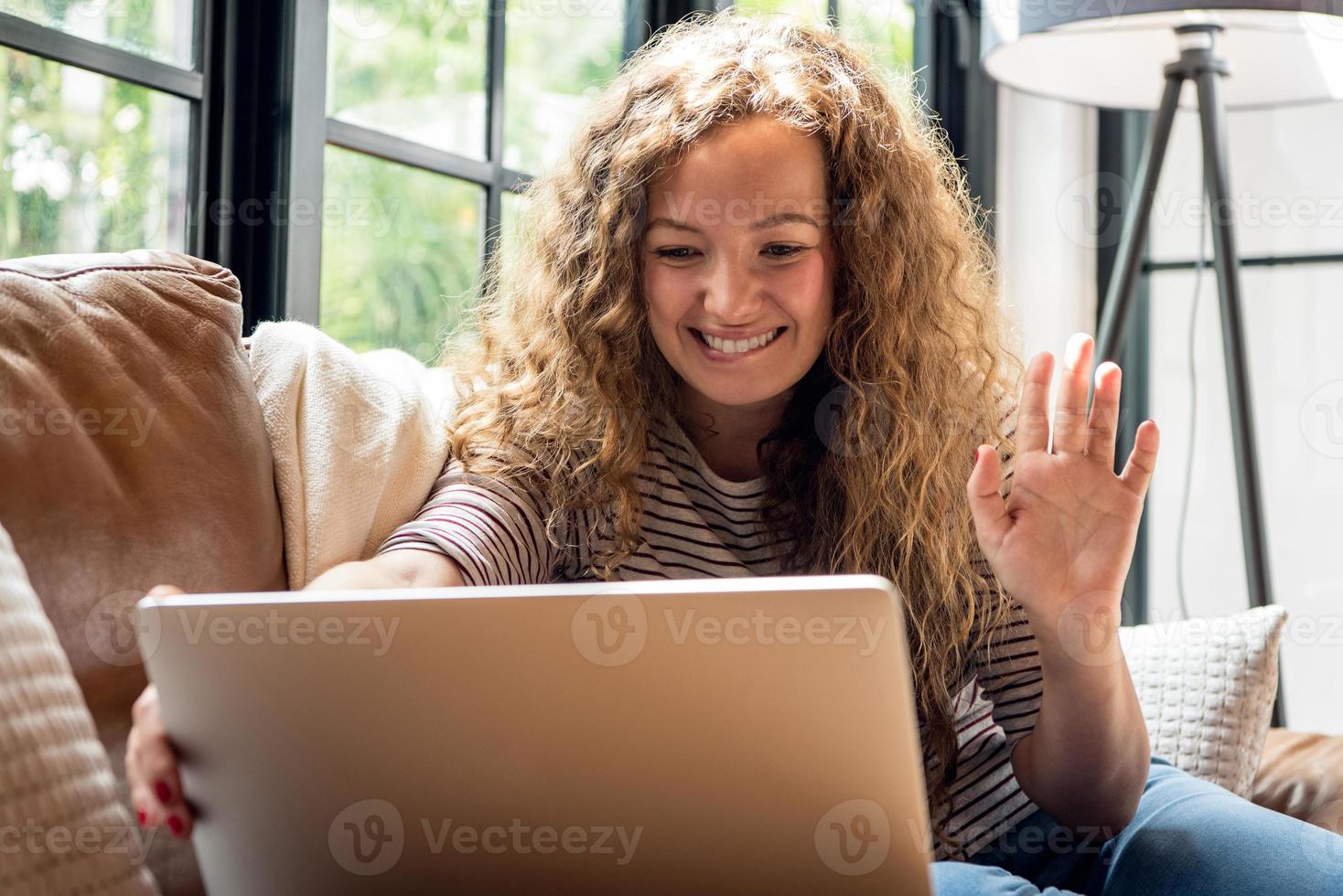 hübsche kaukasische frau, die lächelt und mit der hand zur kamera winkt, während sie einen videoanruf mit einem laptop-computer auf der couch im wohnzimmer zu hause macht foto