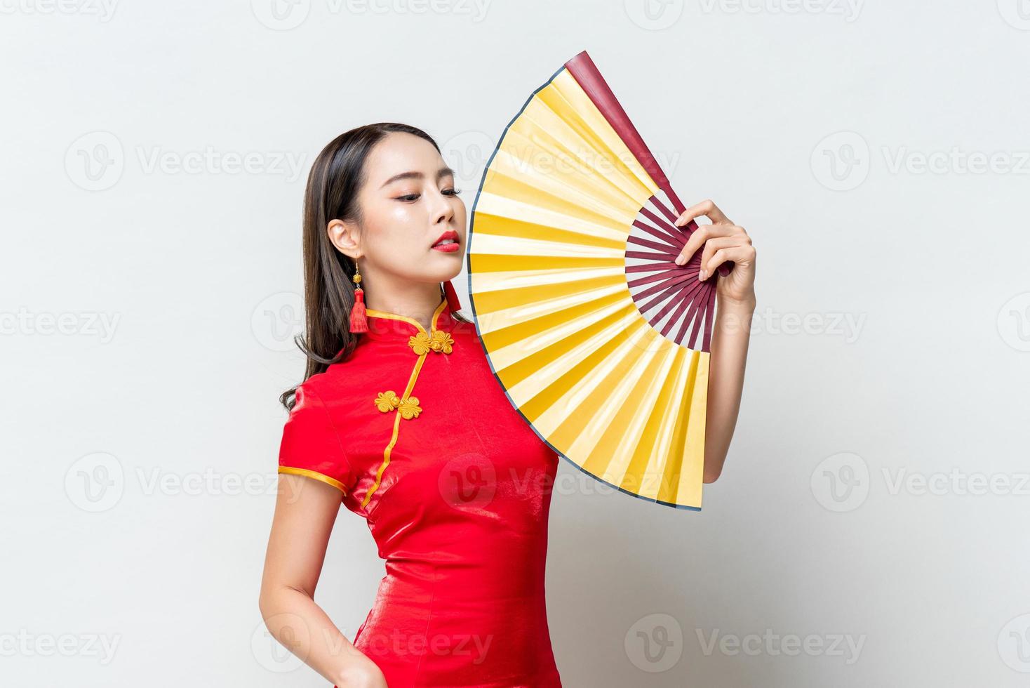 porträt der schönen asiatischen frau, die traditionelles rotes qipao-kleid trägt, das goldenen fan im isolierten hellgrauen hintergrund des studios für chinesisches neujahrskonzept hält foto